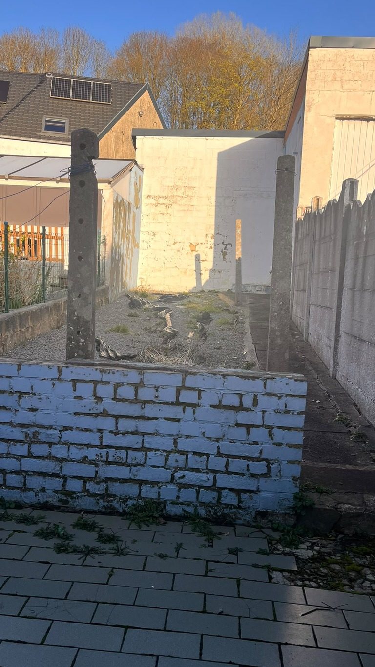 Dezelfde tuin voor de werken: kaal grind met puin en onkruid, betonnen omheiningsplaten en een verweerde muur
